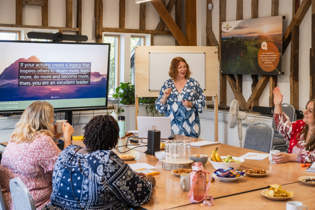 An image of people seated at a meeting room table. There is a speaker at the front, beside a large digital screen.